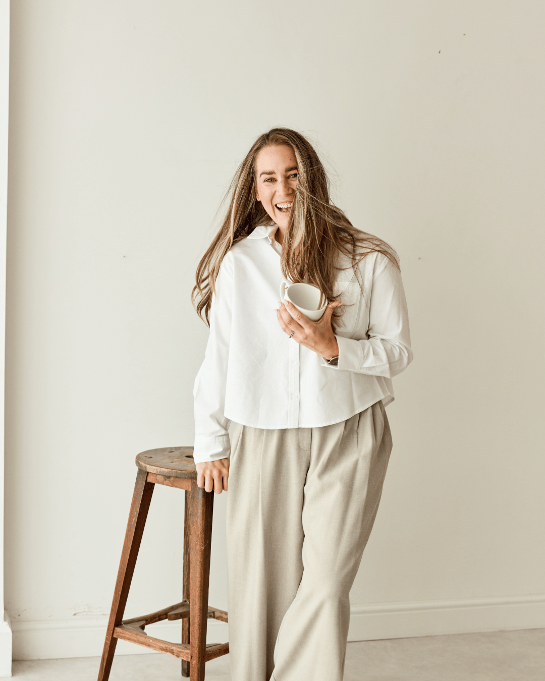 Woman in white shirt and beige pants holding a white mug, standing next to a wooden stool in a bright room, smiling and laughing.
