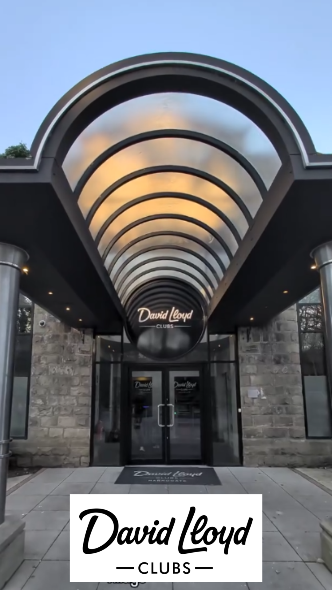 Entrance to David Lloyd Clubs with a glass door and a black round sign. The entrance is covered by a curved glass canopy with black framing. The sign and a mat in front of the door display the David Lloyd logo.
