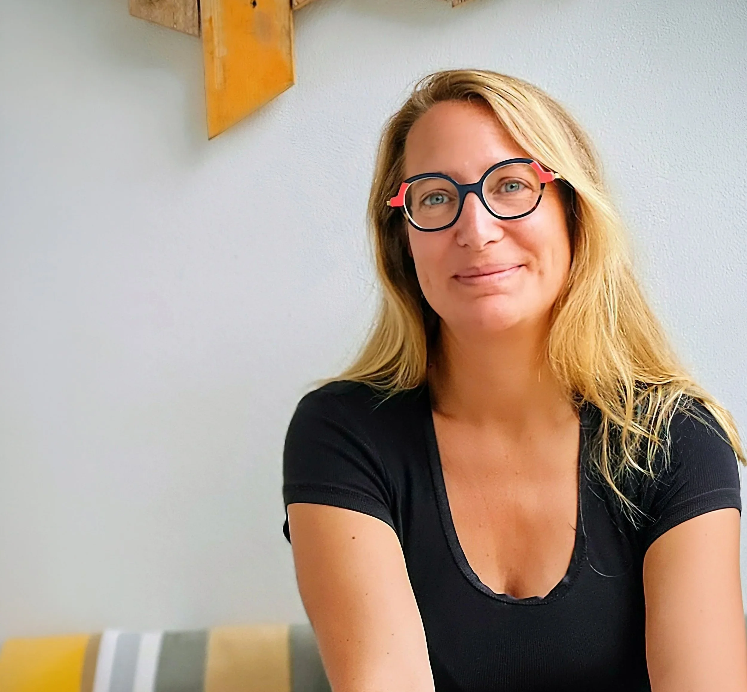 Une femme portant des lunettes noires, souriante, avec des cheveux blonds, près d'un mur blanc avec un tableau en bois