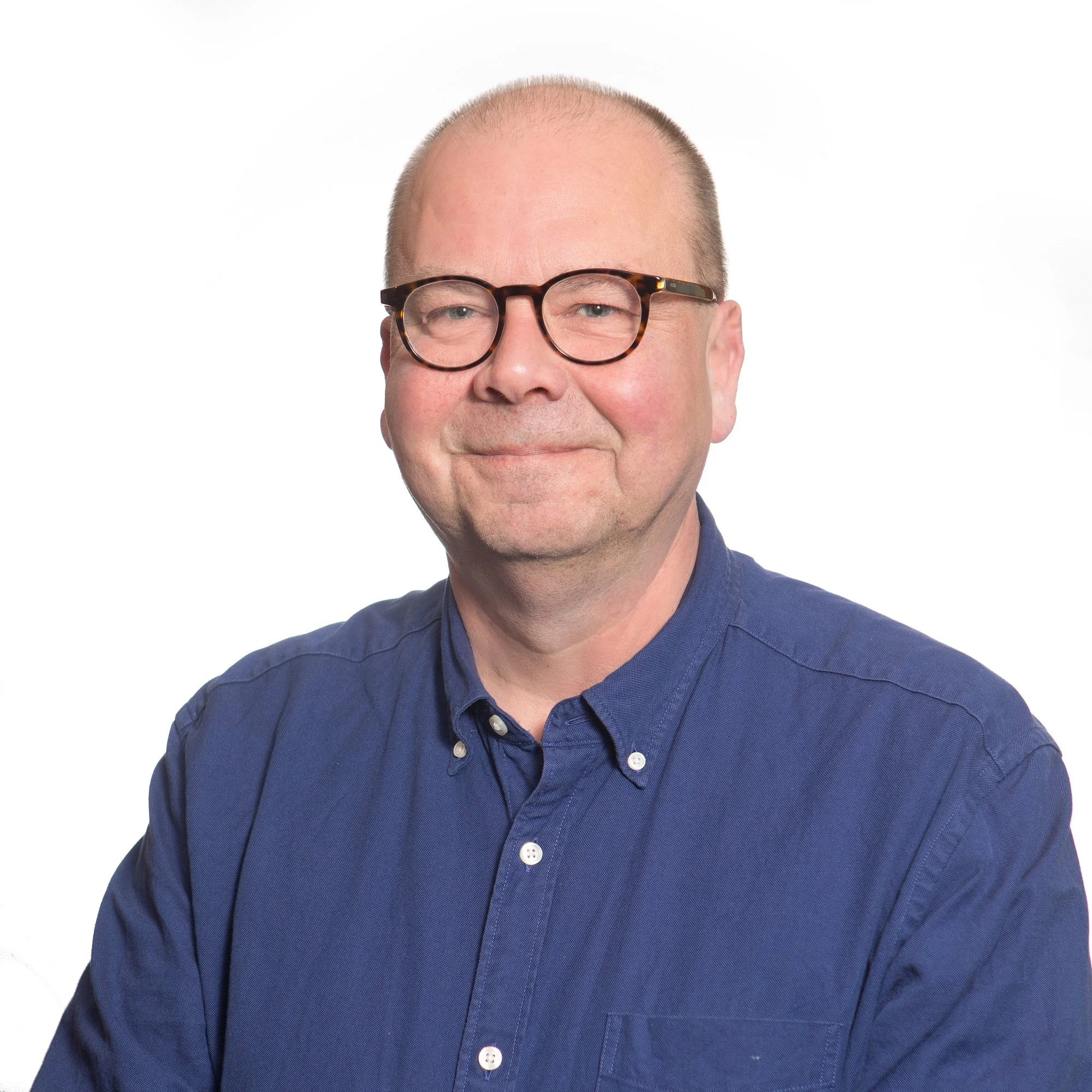 Close-up of a smiling man with short hair, glasses with tortoise-shell frames, wearing a navy blue button-up shirt, against a plain white background.