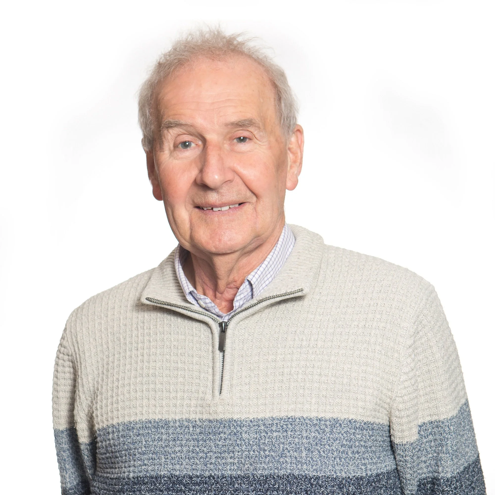 Close-up of an elderly man smiling, wearing a beige zip-up sweater and a checked shirt, against a white background.