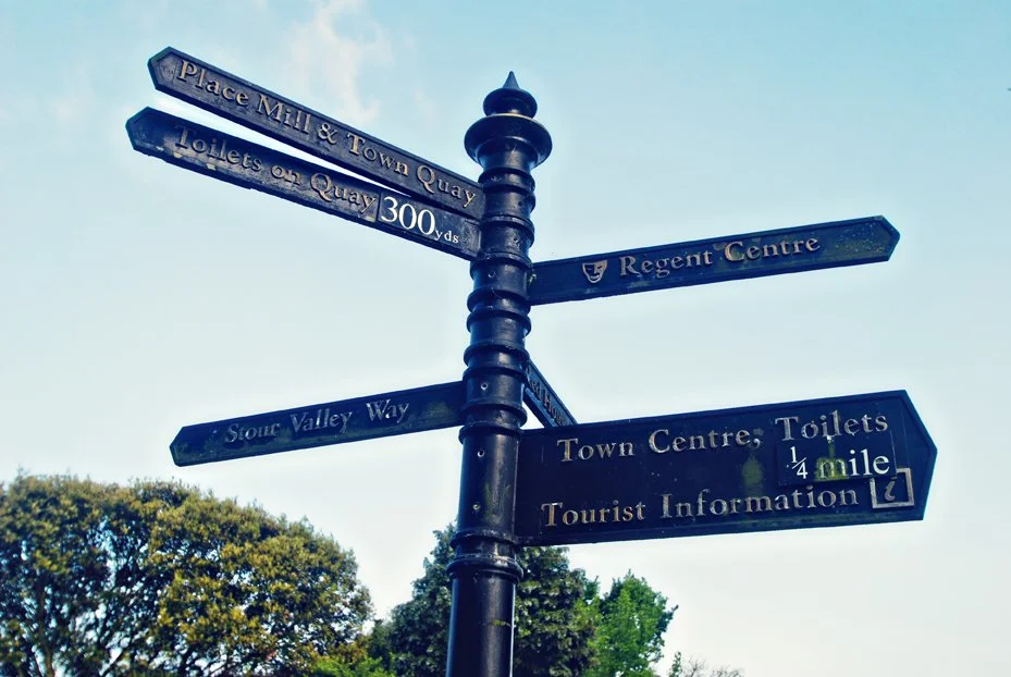 A black street signpost with multiple directional signs pointing to various locations, including the town centre, toilets, Regent Centre, and a tourist information center, with trees and a clear sky in the background.