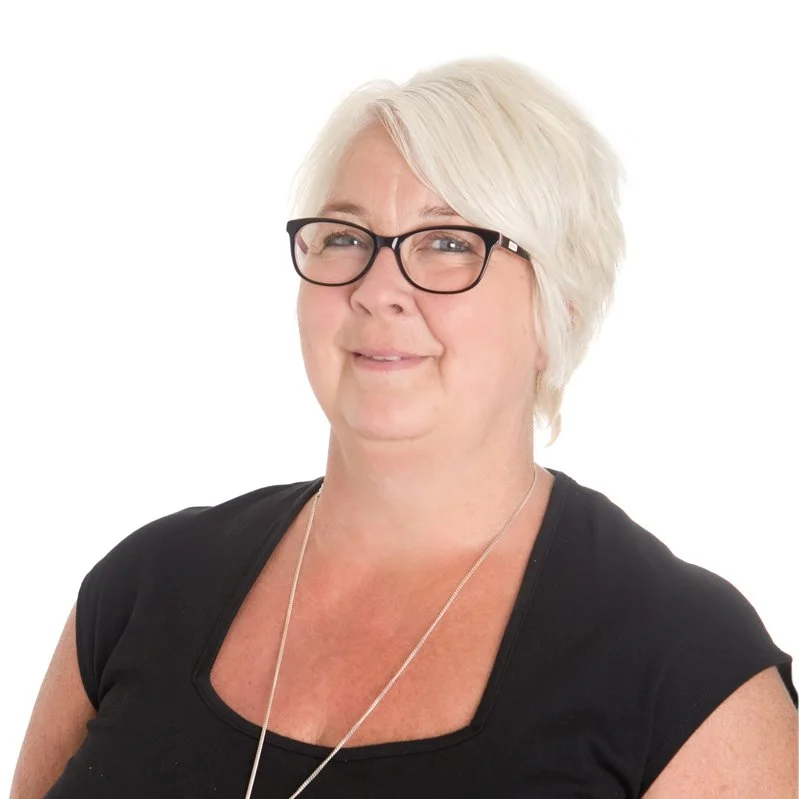A woman with short white hair, wearing black glasses and a black top, smiling at the camera against a white background.