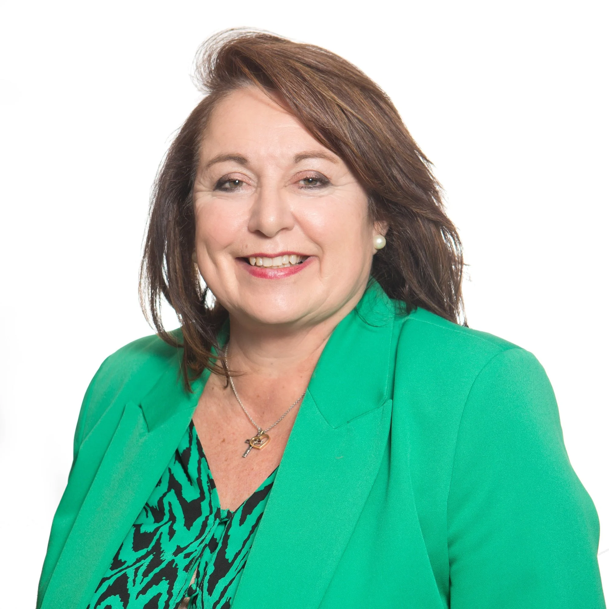 A woman wearing a green blazer and a black and green animal print top, smiling with pearl earrings and a necklace with a heart pendant, with dark brown hair styled in waves.