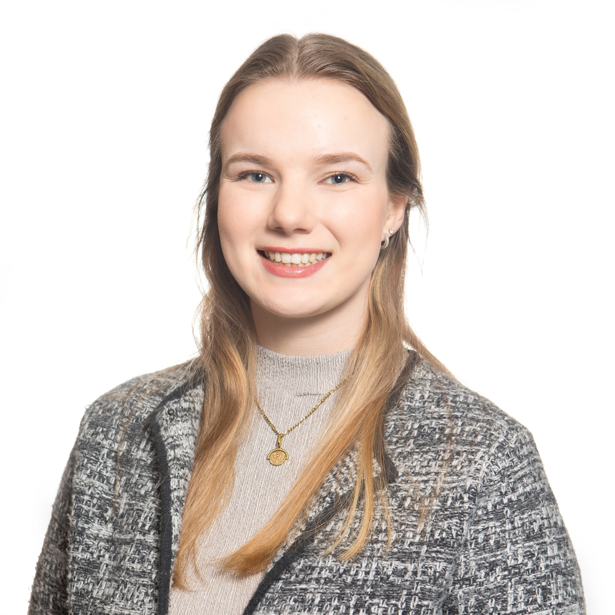 A woman smiling with straight brown hair, wearing a patterned gray blazer, a gray top, and layered gold necklaces, against a plain white background.