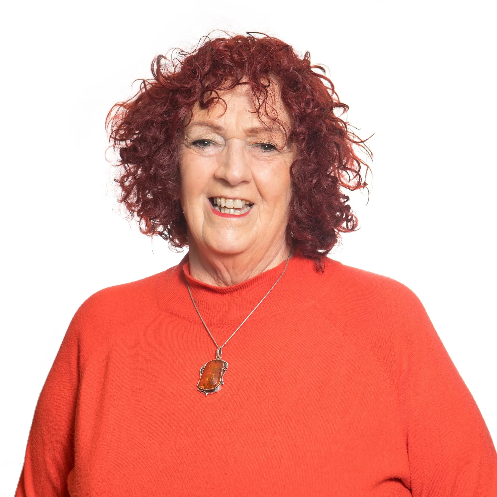 Close-up of a woman with curly red hair, wearing an orange sweater and a pendant necklace, smiling.