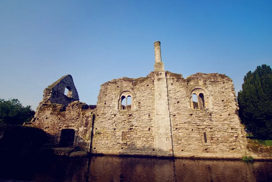 Ruins of an old stone building with a tall chimney, arched windows, and a small doorway, located on the edge of a body of water, under a clear blue sky.