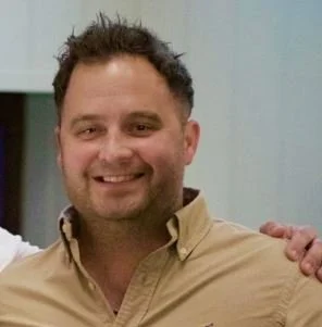 Close-up of a smiling man with short, dark hair, wearing a beige button-up shirt, standing indoors.
