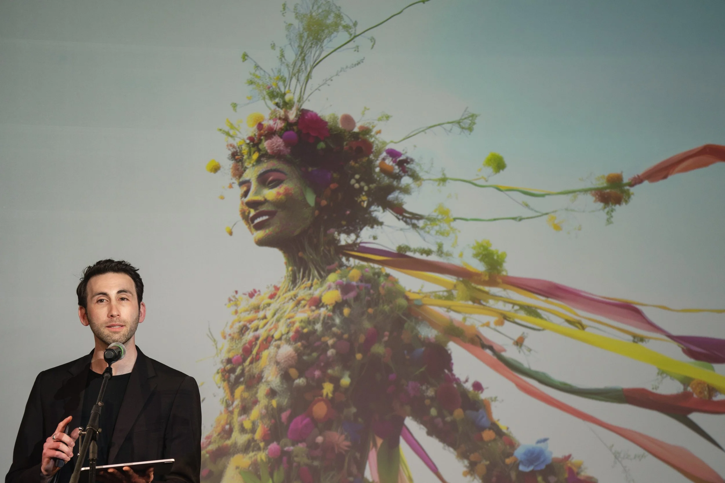 Photograph of a man with dark hair and a beard, wearing a black jacket, speaking into a microphone, with a colorful artistic backdrop of a woman's face made of flowers and paint.