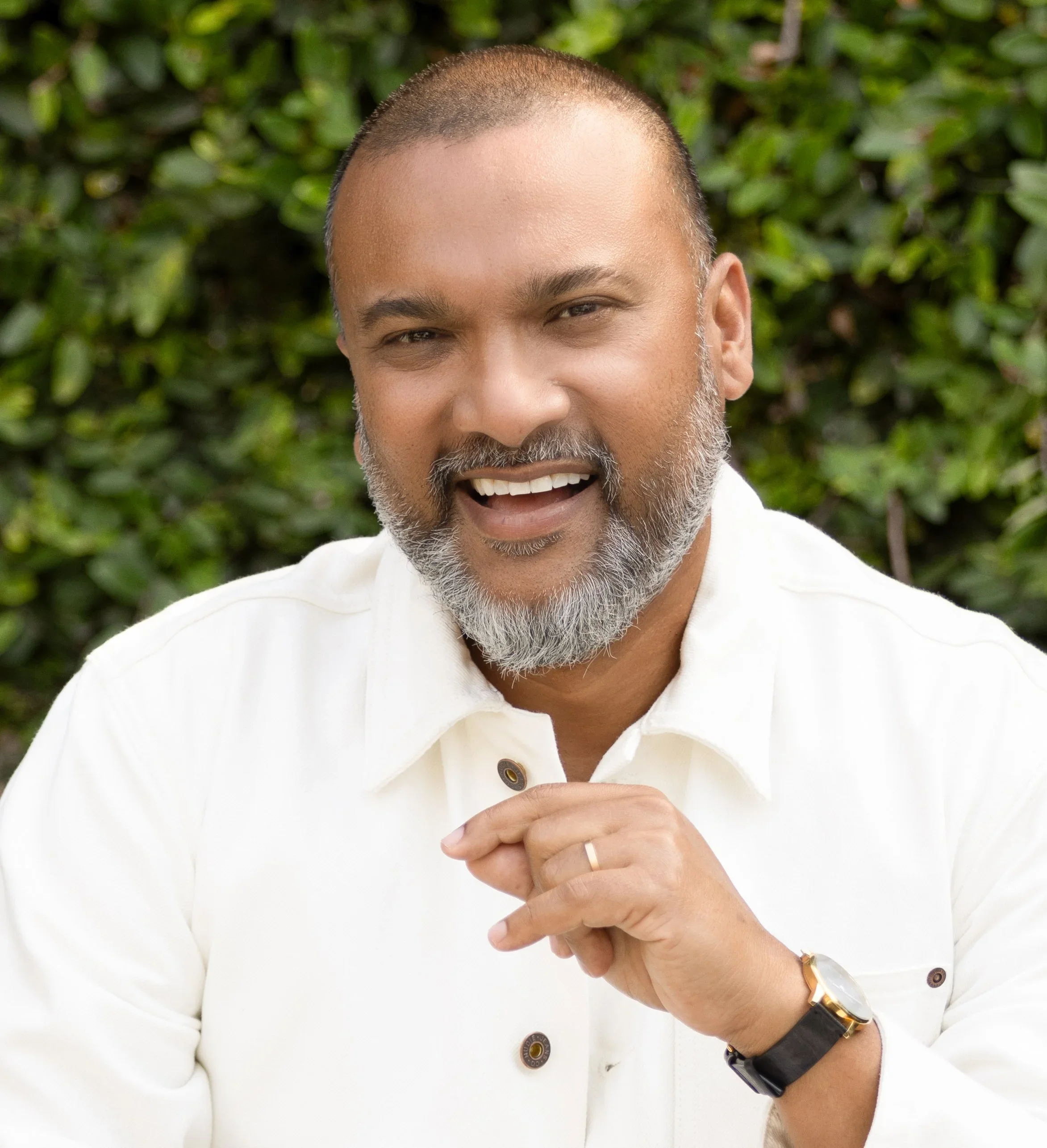 Close-up of a man with short hair and a beard smiling with his mouth open, wearing a white shirt, a black watch, and a gold ring, holding his hand in front of him against a green leafy background.