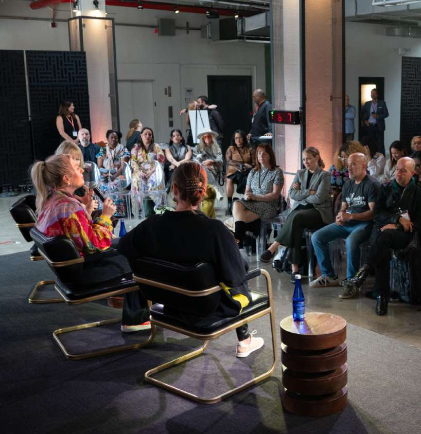 A panel discussion with two women speaking at the front of a room, facing an audience of about 20 people. The setting appears to be a conference or seminar, with a camera crew and audience members listening attentively. The room has black and white decor and a digital clock showing 6:11.