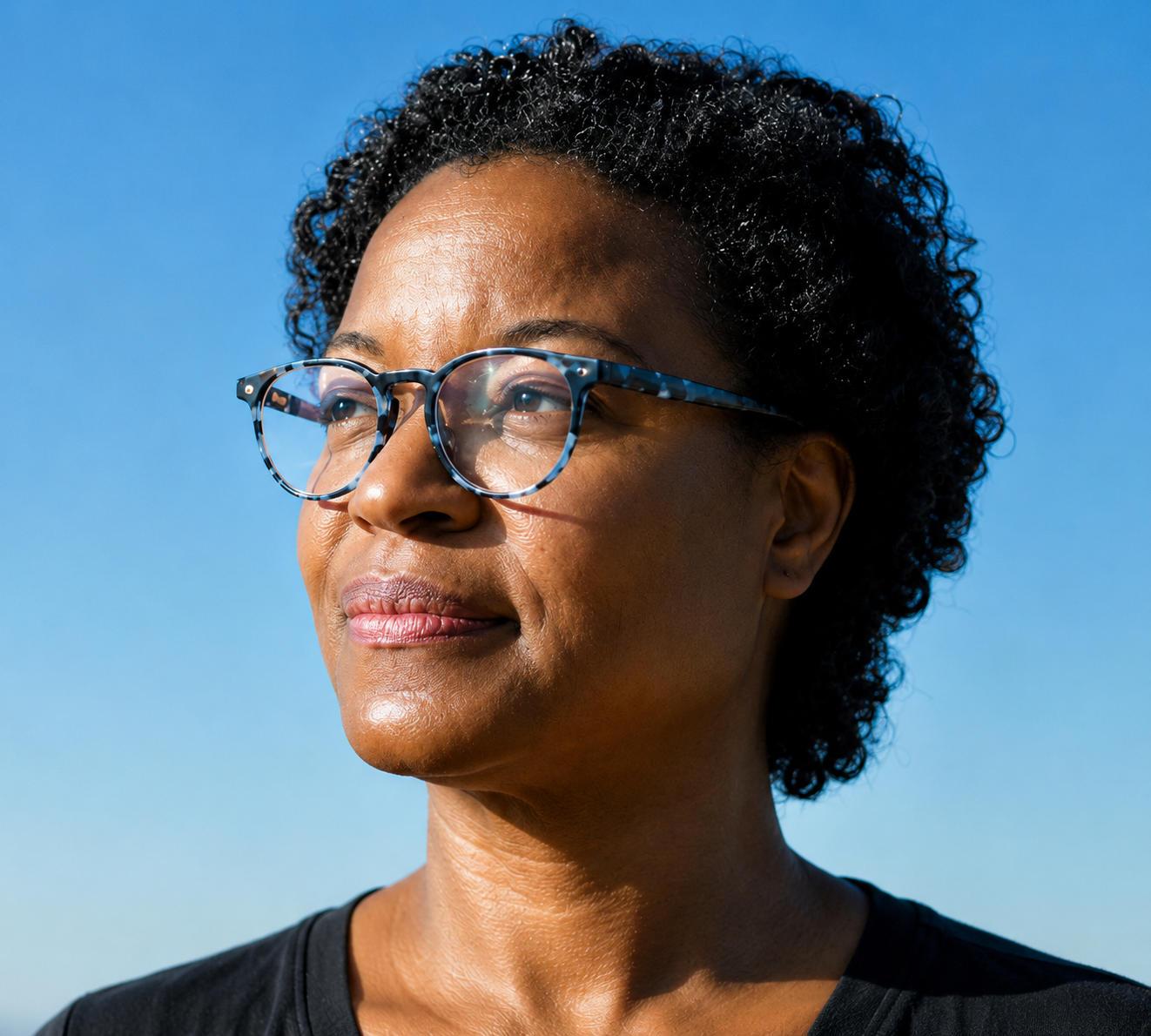 A middle-aged woman with short curly black hair and glasses looking into the distance against a clear blue sky.
