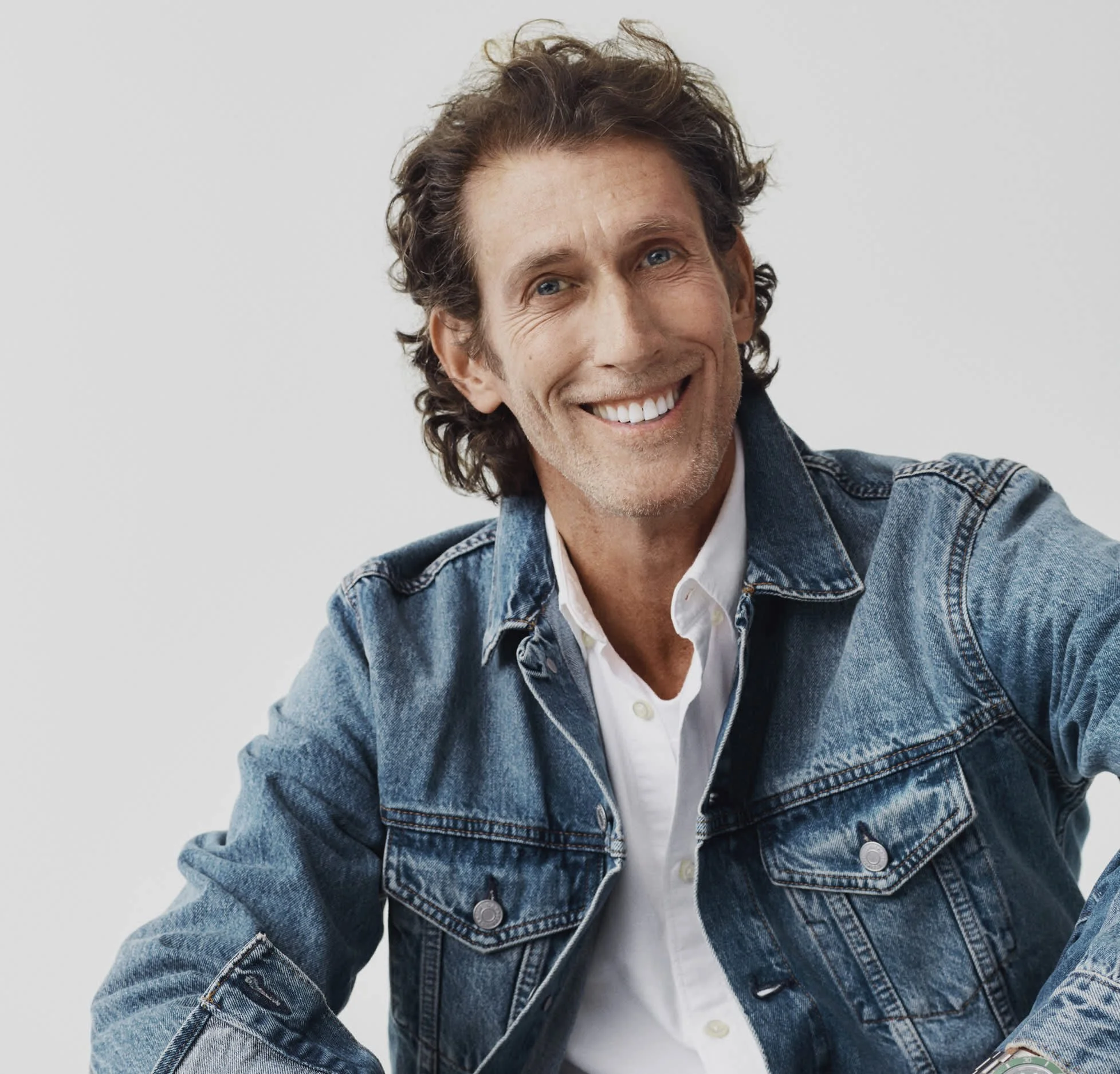 A close-up of a smiling man with wavy brown hair, blue eyes, wearing a denim jacket over a white shirt, against a plain light background.