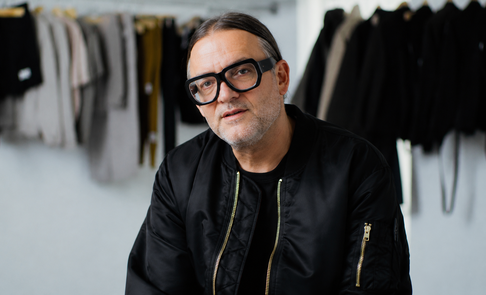 A middle-aged man with gray beard and glasses, wearing a black jacket, sitting in a room with clothing hanging on racks in the background.