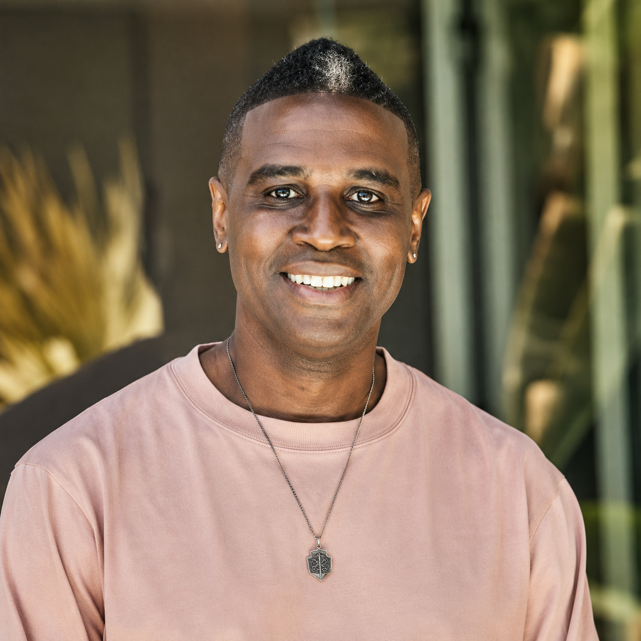 A smiling man with earrings, wearing a pink shirt and a necklace with a pendant, standing outdoors with blurred green foliage in the background.