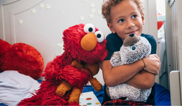 A young boy hugging a stuffed animal with a Sesame Street character plush toy beside him in a cozy room.