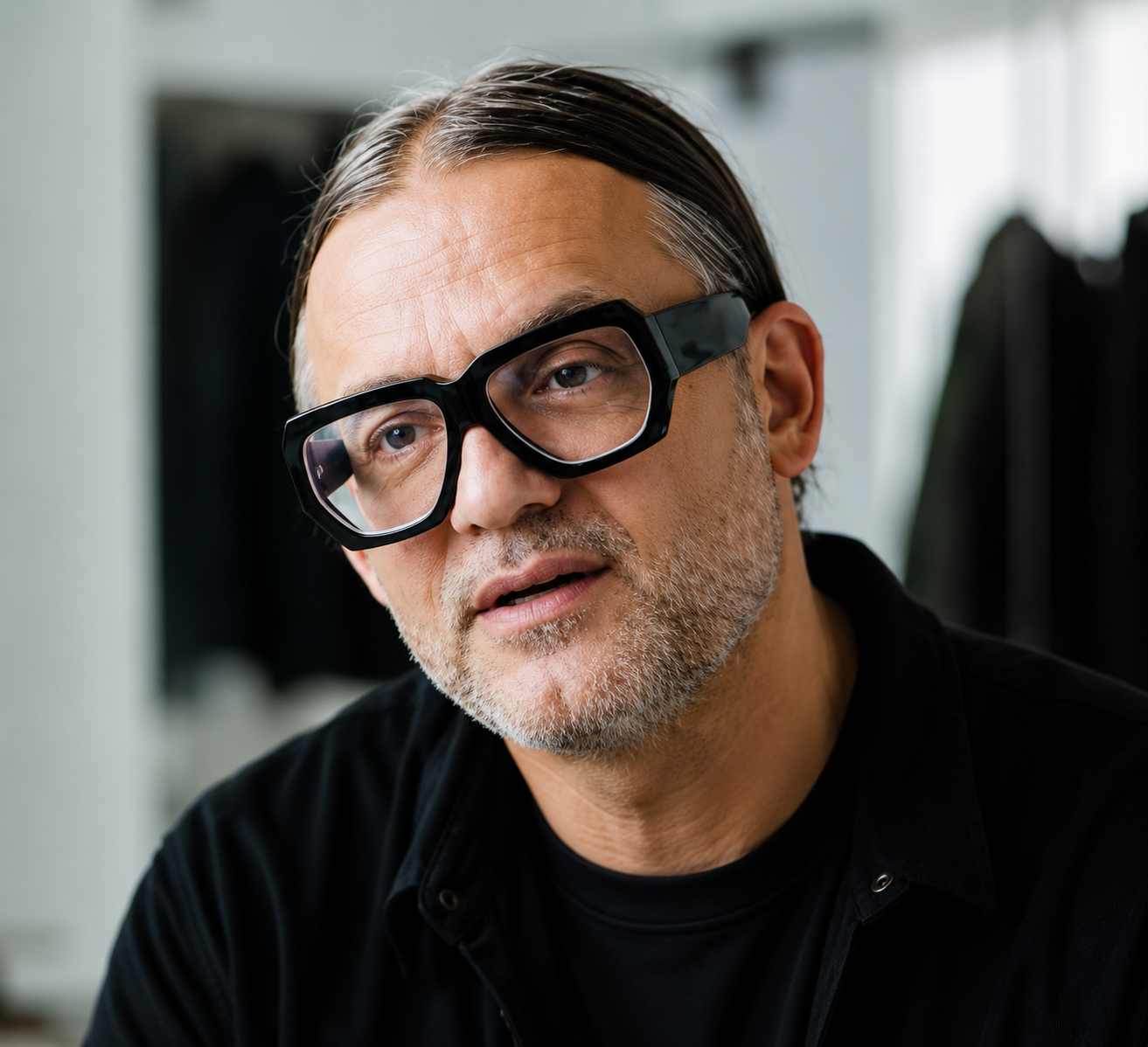 A middle-aged man with gray hair and beard wearing black, oversized glasses, looking slightly to the side.