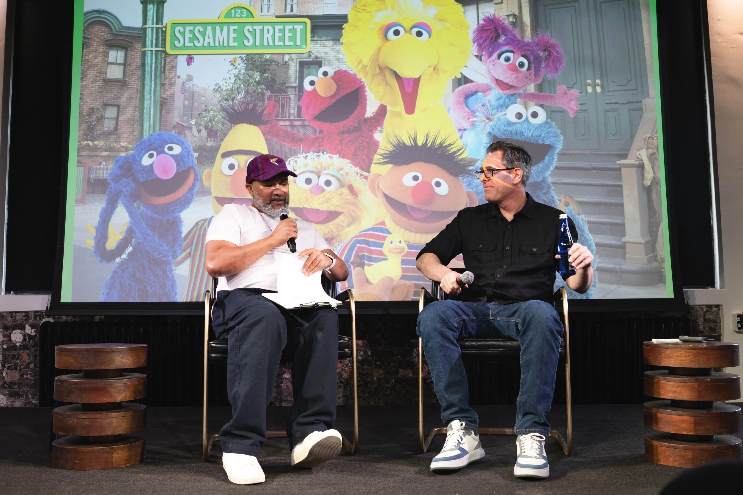 Two men are seated on stage in front of a large colorful backdrop of Sesame Street characters. The man on the left is wearing a purple cap and a white T-shirt, holding a microphone. The man on the right is wearing glasses, a black shirt, and blue jeans, holding a water bottle. The backdrop features characters like Big Bird, Elmo, Zoe, Telly, and others.