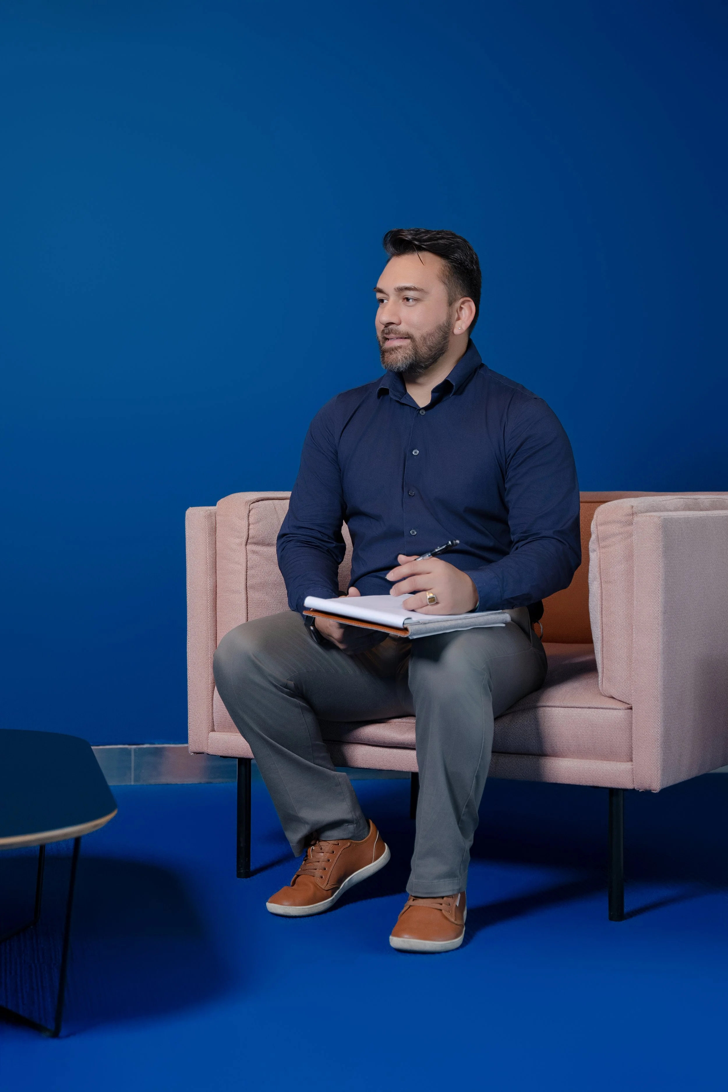 A man with a beard in a blue shirt sitting on a pink sofa against a blue background, holding a notebook and pen.