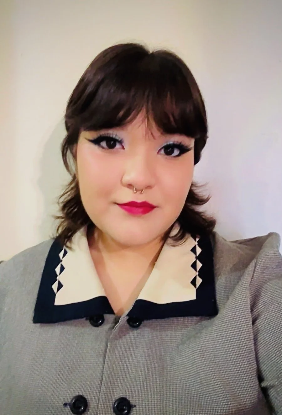 A woman with short, dark hair, wearing makeup with bold eyeliner and red lipstick, a gray blazer with a patterned collar, and a septum piercing, posing for a selfie against a plain background.