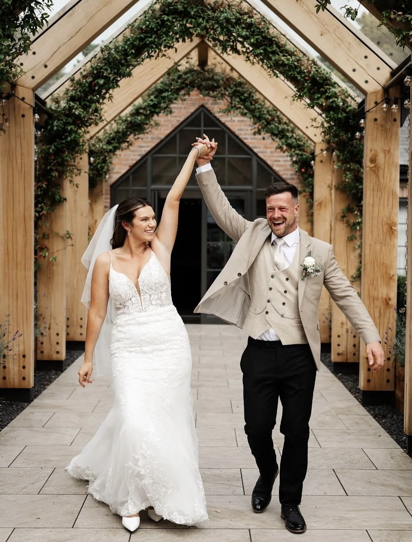 A bride and groom celebrating their wedding outdoors under a wooden arch decorated with greenery, smiling and holding hands up in victory.