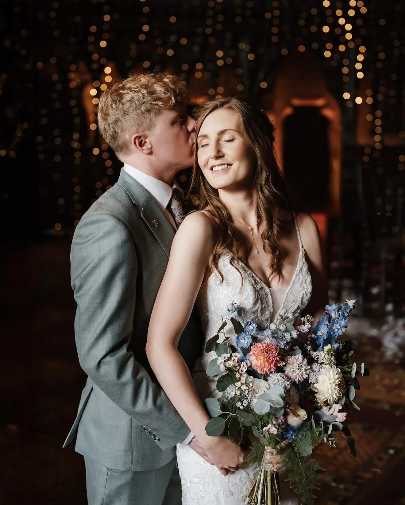 A bride and groom share a tender moment during their wedding celebration. The groom gently kisses the bride's temple while she smiles with her eyes closed, holding a large bouquet of colorful flowers. The background features warm, blurry lights, crea
