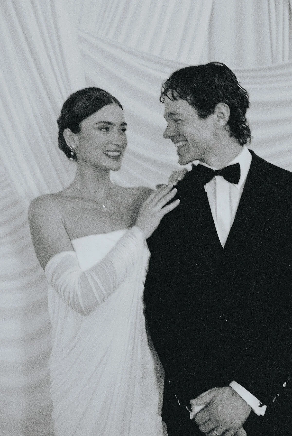 A bride and groom in wedding attire share a joyful moment, smiling at each other against a draped fabric background.