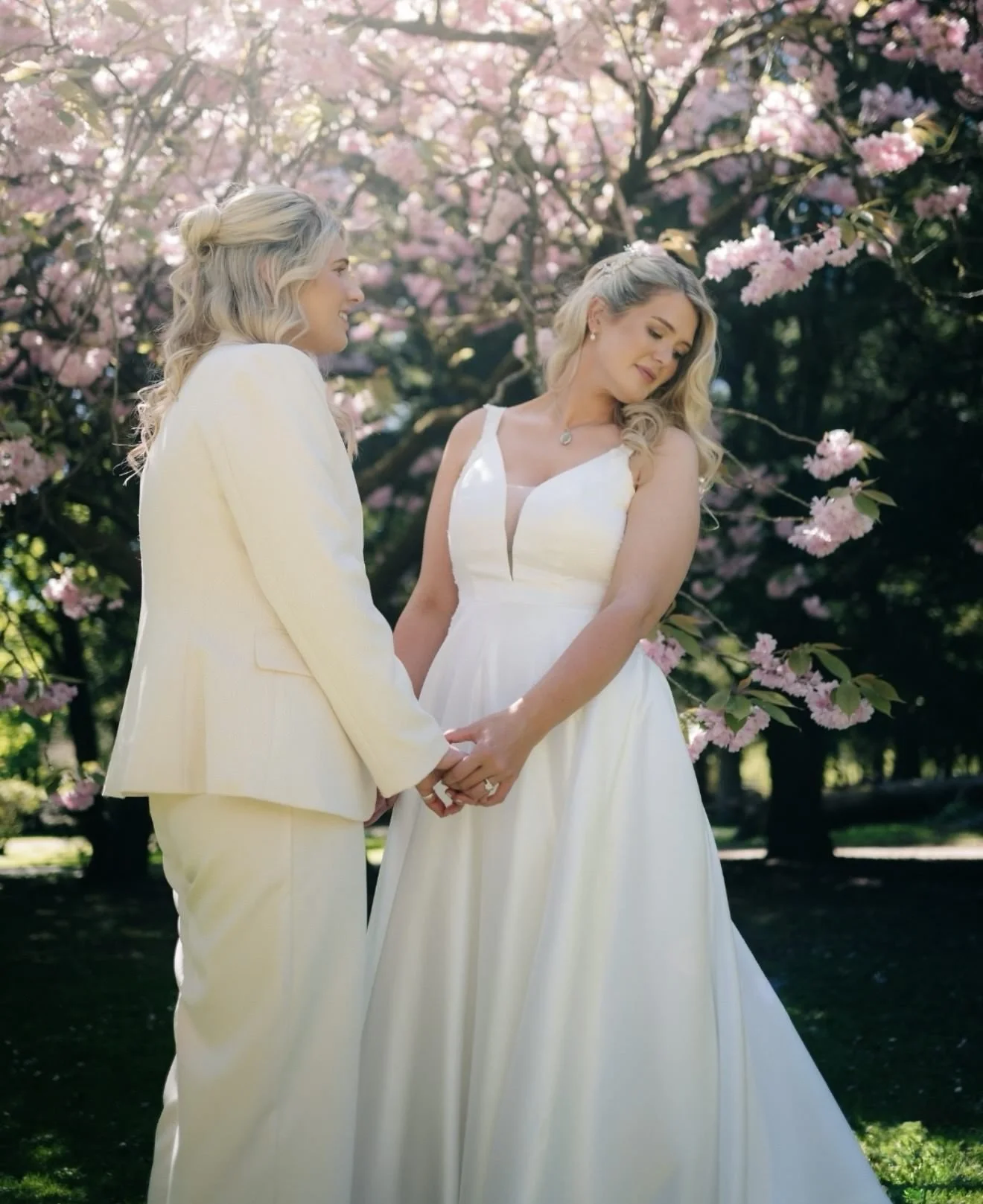 Two women holding hands, standing outdoors in front of pink flowering trees, dressed in white wedding attire, during a wedding or commitment ceremony.