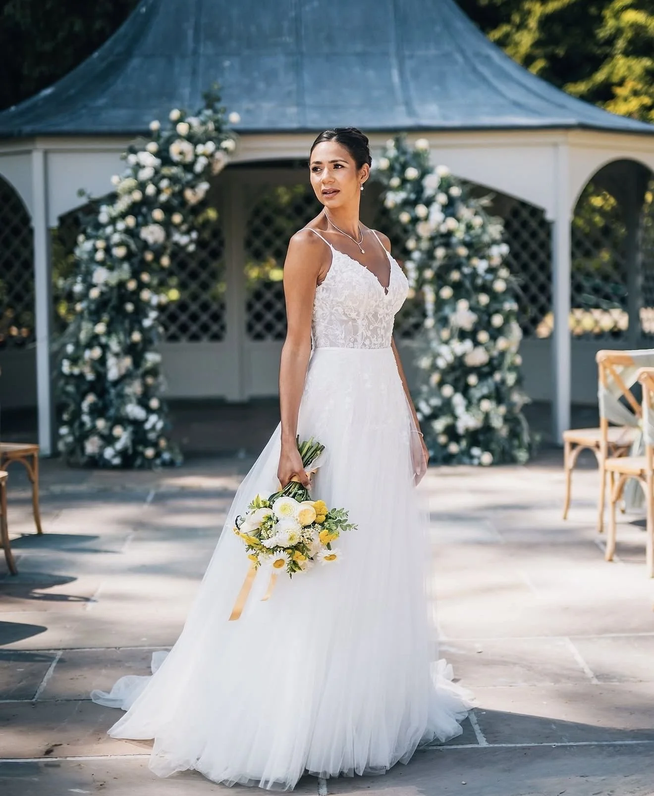 A bride in a white wedding gown holding a bouquet stands outdoors in front of a decorated gazebo during daylight.