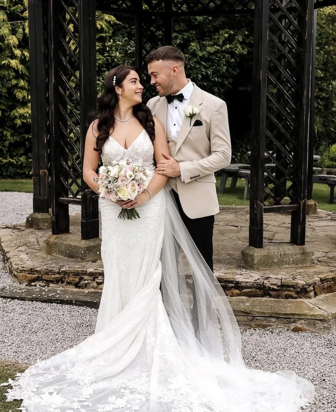 A newlywed couple on their wedding day, standing outdoors under a wooden arch, with the bride holding a bouquet of pink and white flowers, both smiling and gazing at each other.