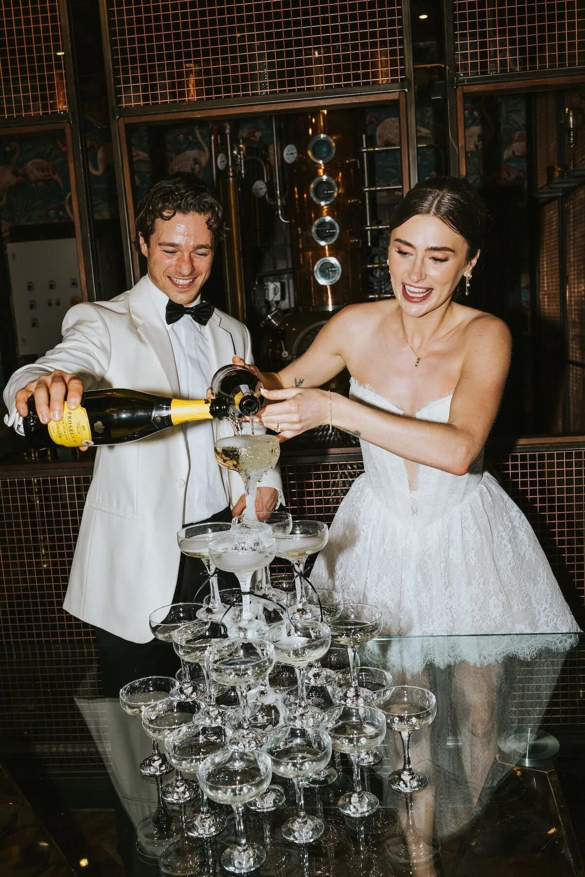 A bride and groom in wedding attire pouring champagne into a tower of glasses during their celebration.