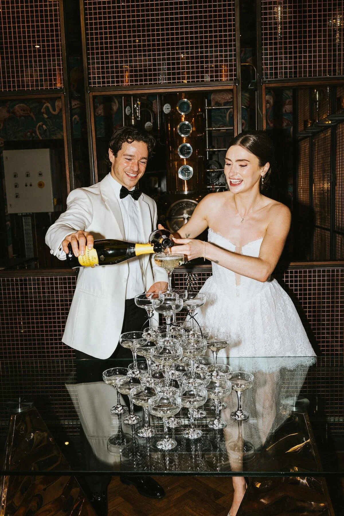 A couple in wedding attire pouring champagne into a tower of glasses at their wedding reception. The groom is wearing a white tuxedo jacket and the bride is in a white wedding dress, both smiling.