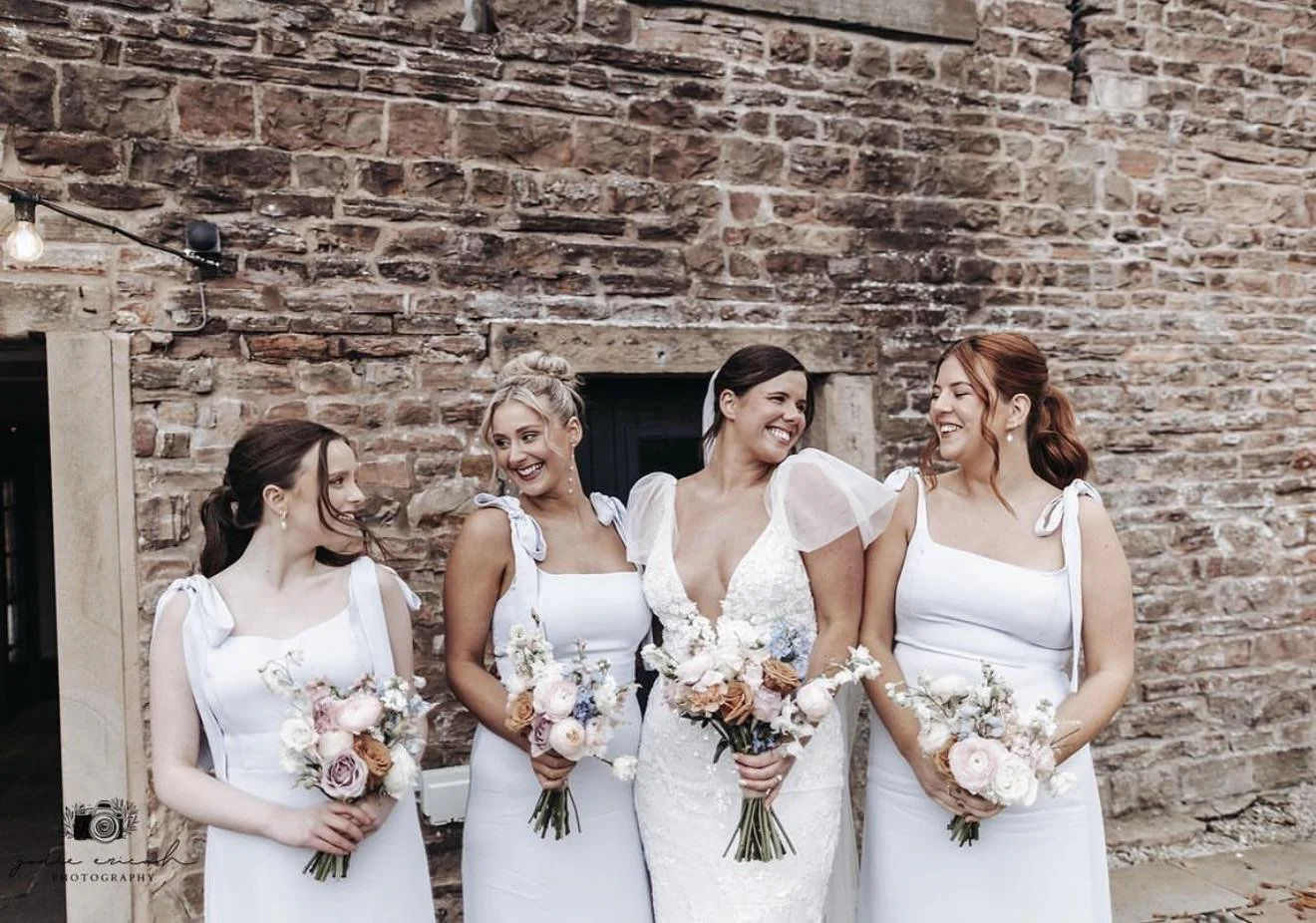 Four women in white dresses holding flowers, smiling and looking at each other, standing against a brick wall.