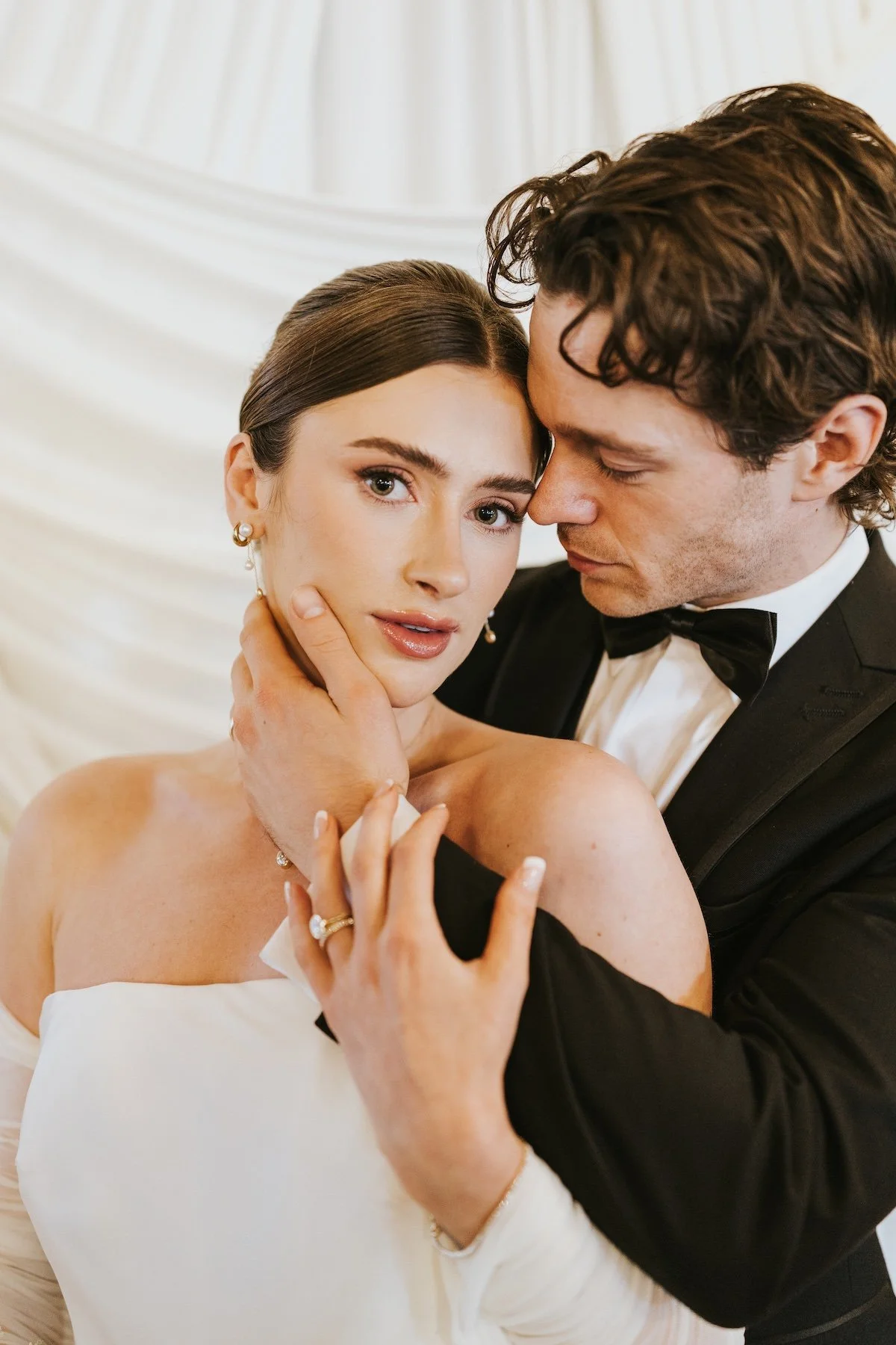 A bride and groom in a tender moment, with the bride looking directly at the camera and the groom gently touching her face, both dressed in wedding attire.