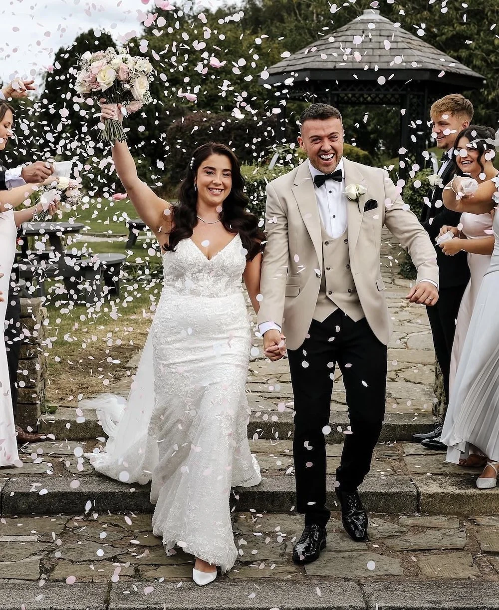 A newlywed couple walking hand-in-hand outdoors, celebrating their wedding with confetti and friends. The bride holds a bouquet and smiles brightly, while the groom wears a beige tuxedo with a black bow tie, both surrounded by well-wishers.