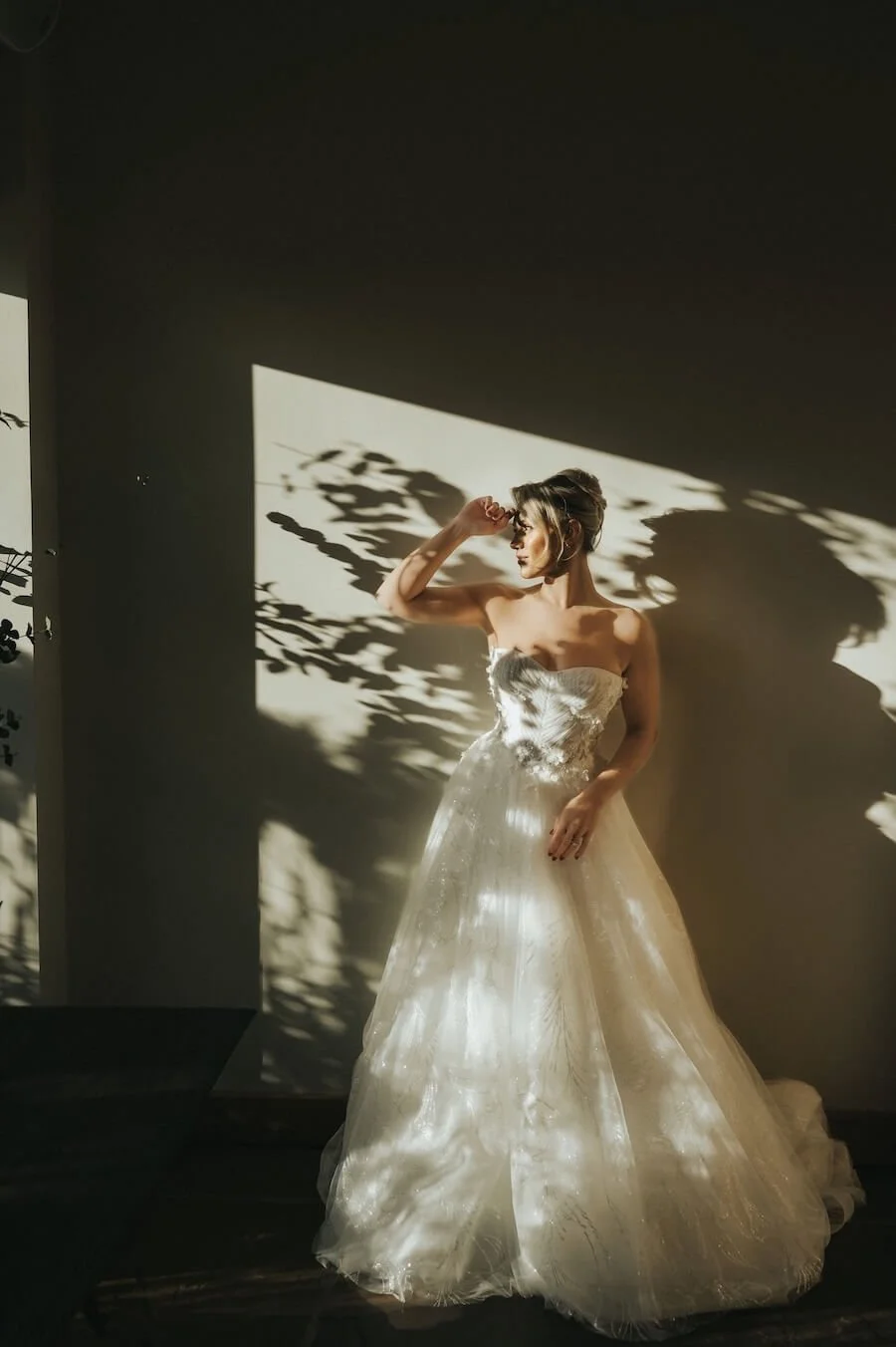 Bride in a strapless wedding dress standing in front of a wall casting shadows of leaves, with sunlight creating patterns on her dress and background.