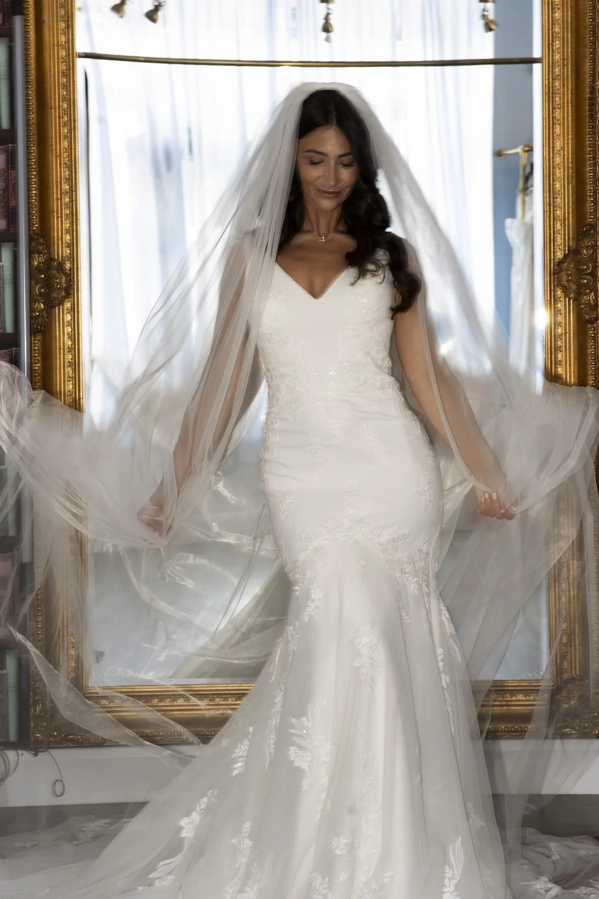 A woman in a white wedding gown and veil standing in front of a large, ornate gold framed mirror, with light-colored curtains in the background.