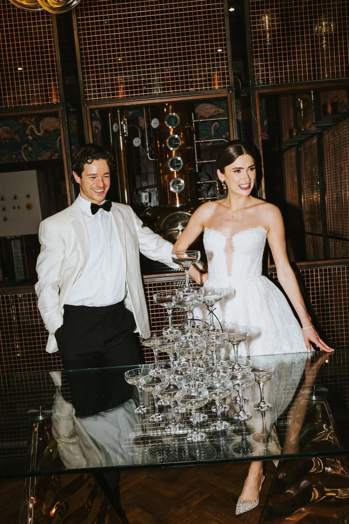 A couple in wedding attire celebrating with a tower of champagne glasses in a stylish, dimly lit setting.