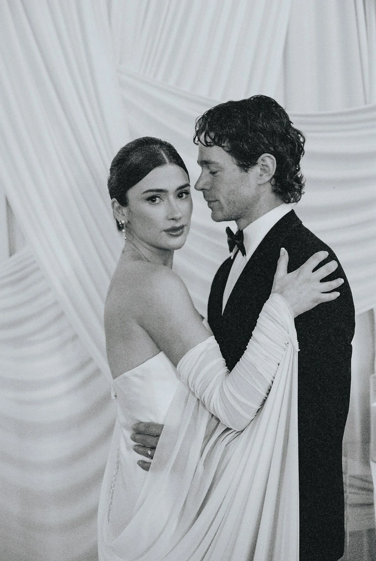 A black-and-white photo of a bride and groom, closely embracing, with a draped backdrop. The bride is wearing an off-shoulder wedding gown with gloves, and the groom is dressed in a tuxedo with a bow tie.