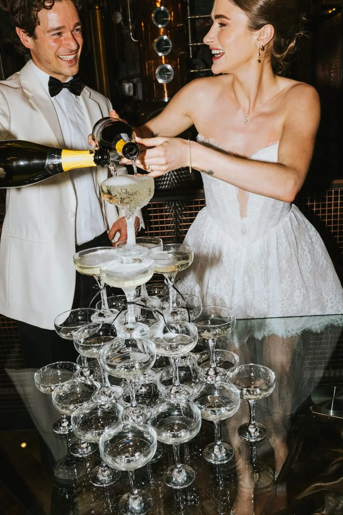 A bride and groom in wedding attire celebrate as they pour champagne into a champagne tower at their wedding reception.