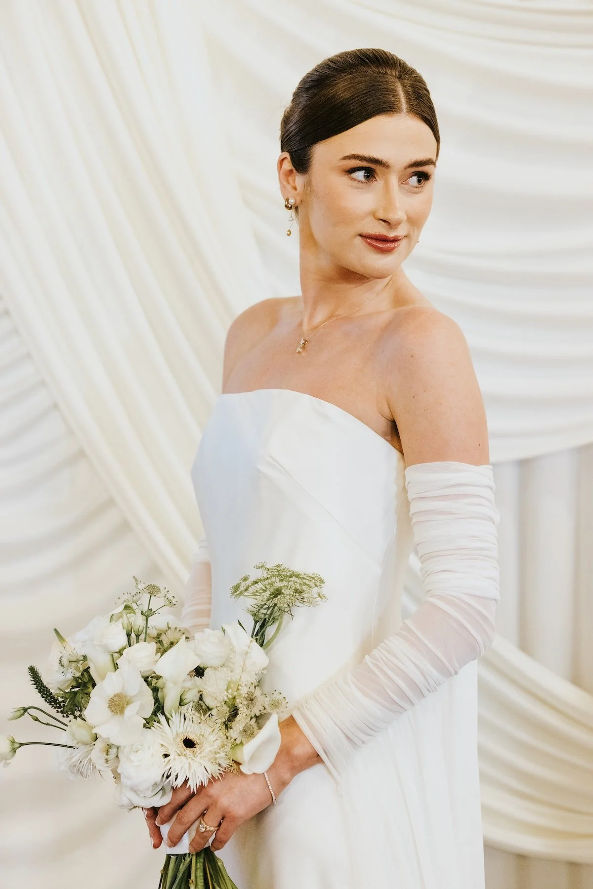 A bride holding a bouquet of white flowers, wearing a strapless white wedding dress with long sheer gloves, and smiling softly.