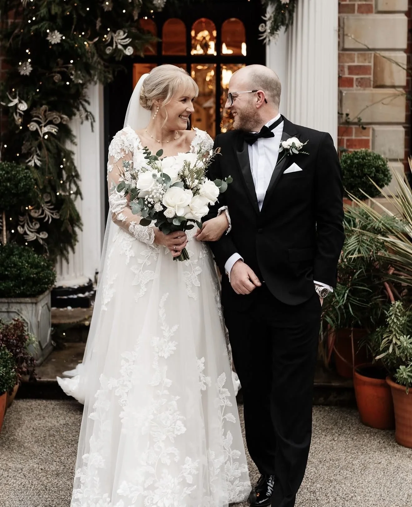 A bride in a lace wedding gown holding a bouquet of white flowers and greenery, smiling at the groom. The groom, dressed in a black tuxedo with a bow tie and glasses, is smiling back. They are standing outdoors near a building with brick and column a