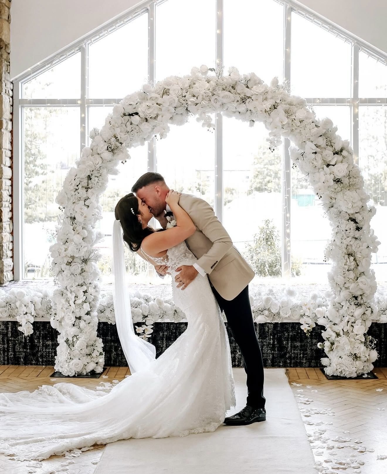 A bride and groom sharing a kiss during a wedding ceremony, standing under a white floral arch in a bright indoor venue with large windows.