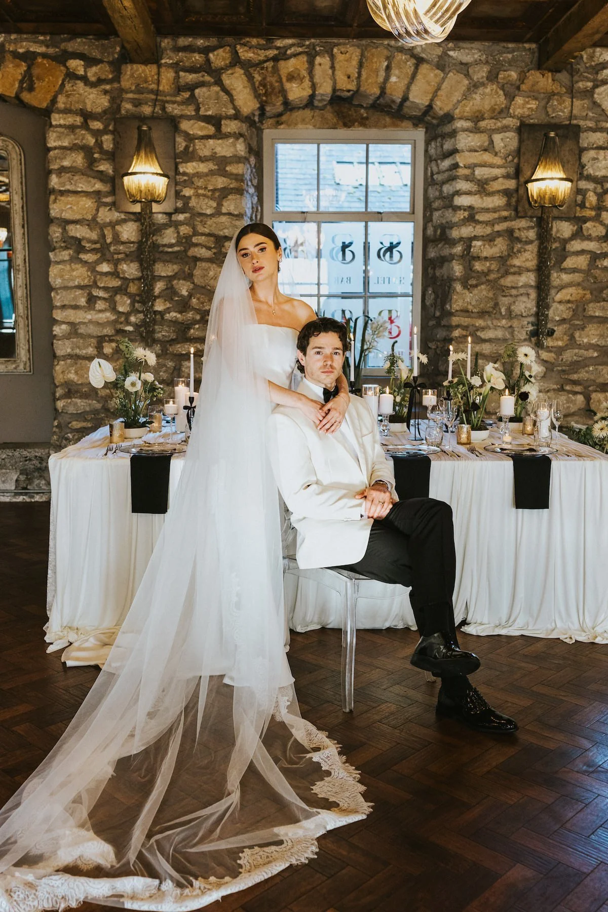 A bride in a white wedding dress and veil standing next to a groom in a tuxedo with a white jacket, seated at a decorated reception table in a rustic stone-walled venue.