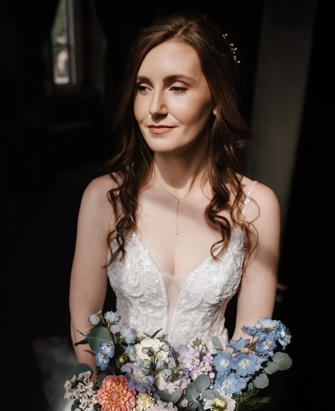 A bride with long auburn hair in a white lace wedding dress, holding a colorful bouquet of flowers, in a dimly lit room