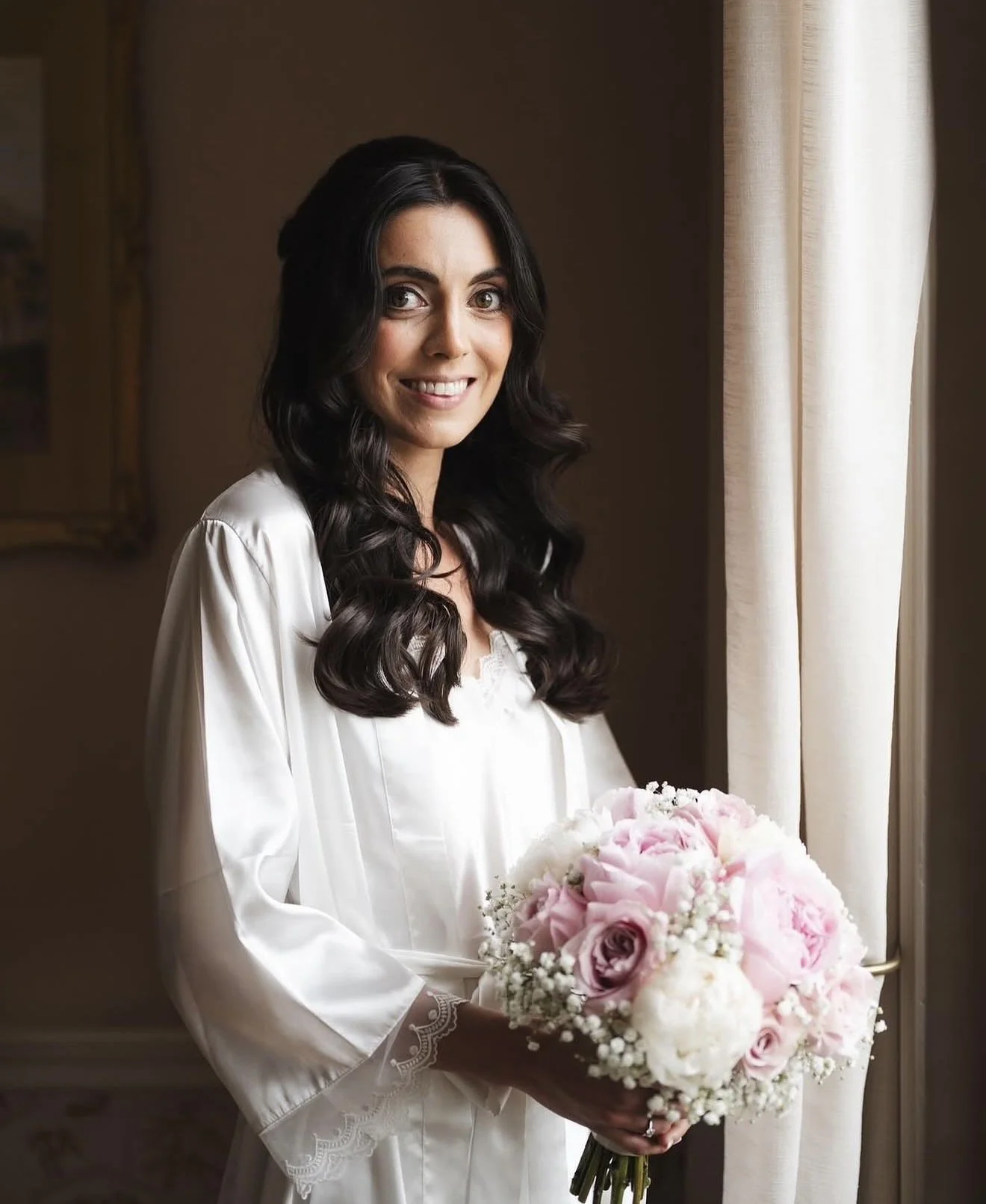 A woman with dark wavy hair smiling while holding a bouquet of pink and white flowers, standing near a window with cream-colored curtains.