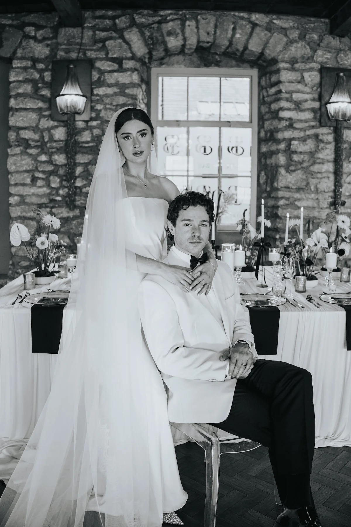 Black and white photo of a bride and groom at a wedding reception. The bride, wearing a strapless wedding gown and a veil, is standing behind the seated groom, who is dressed in a tuxedo with a bow tie. The setting includes a decorated dining table w