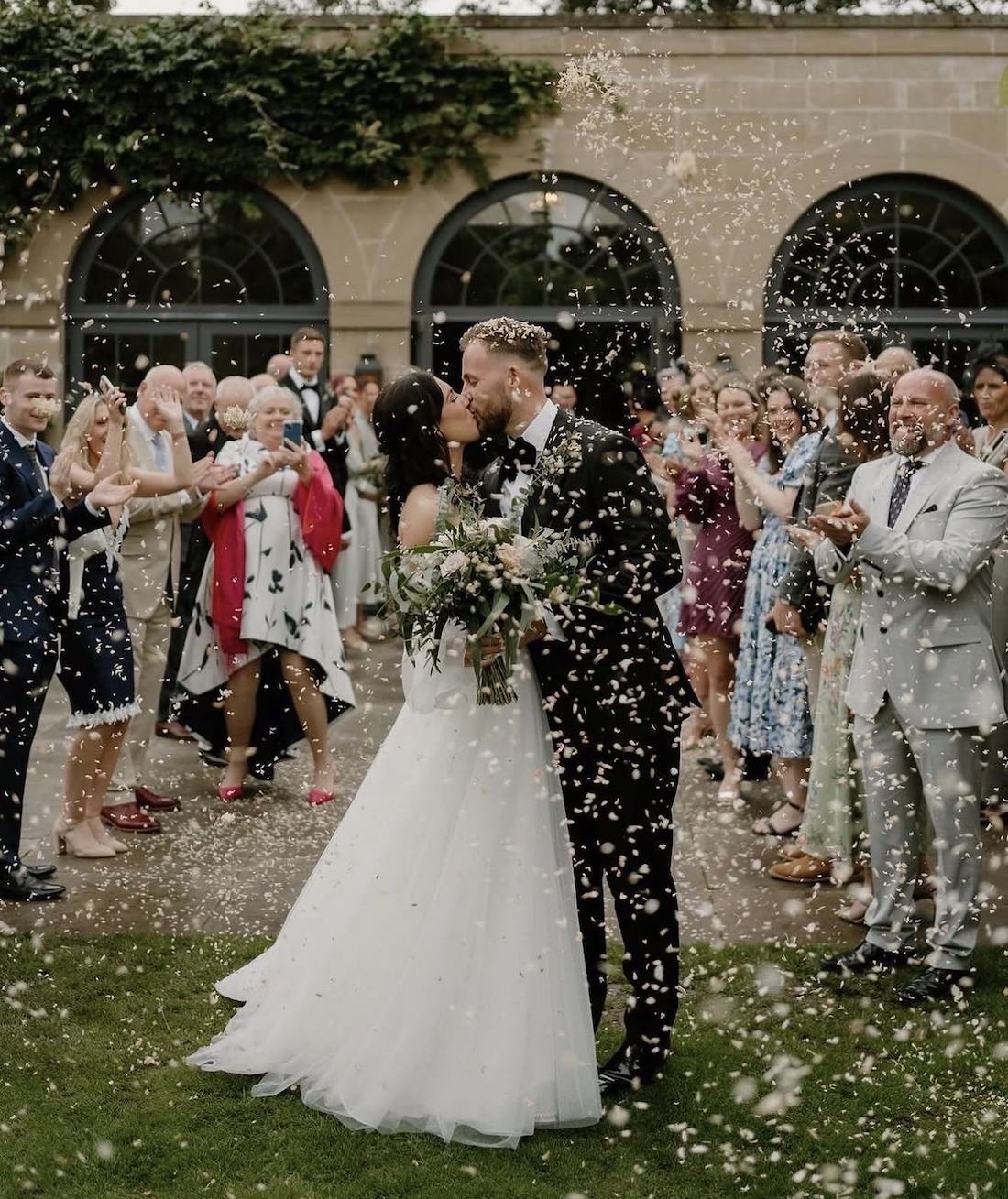 Newlyweds kissing under falling confetti during a wedding celebration with guests clapping and taking photos.