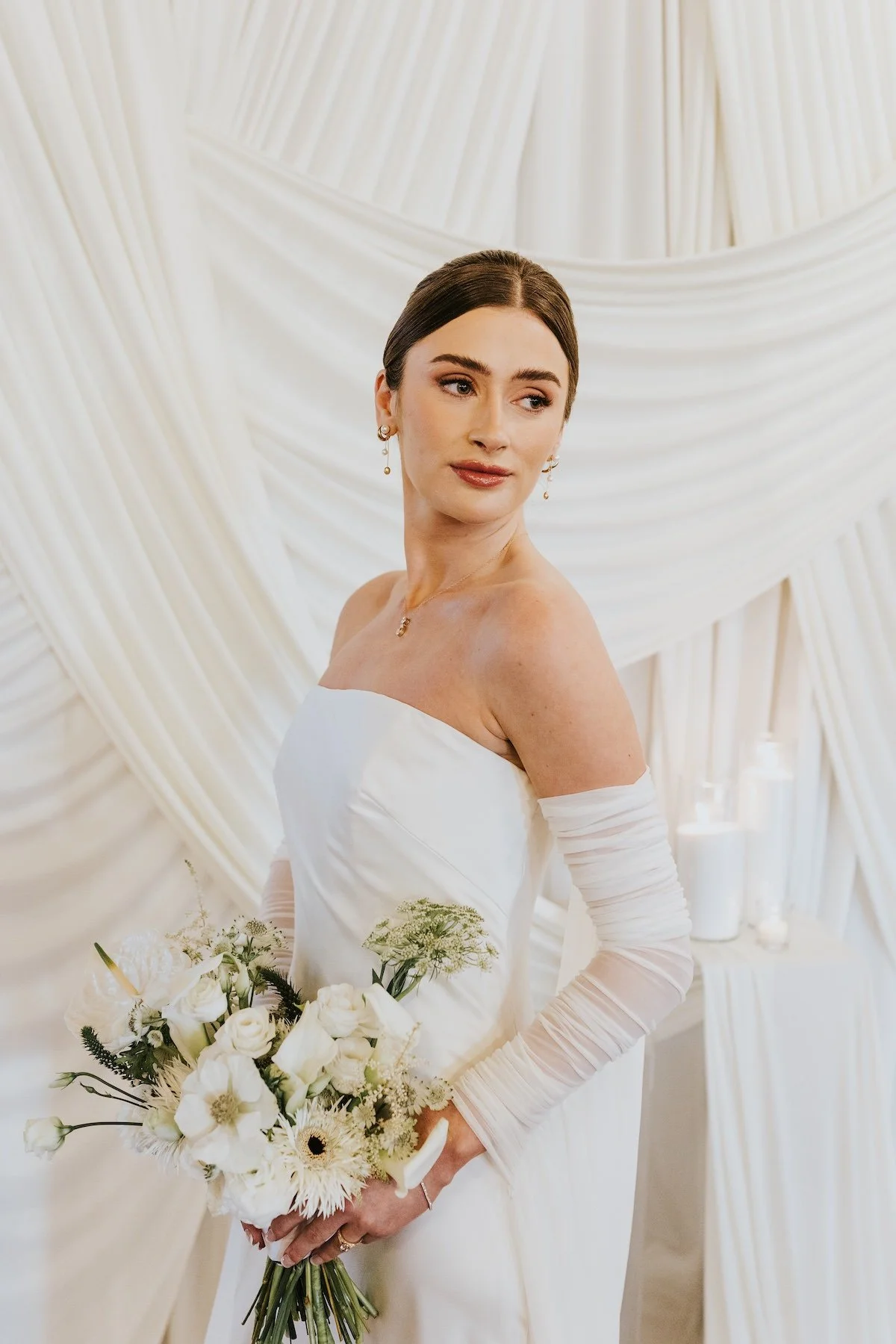 A bride in a white strapless wedding dress holding a bouquet of white flowers, standing in front of a cream-colored draped fabric backdrop.