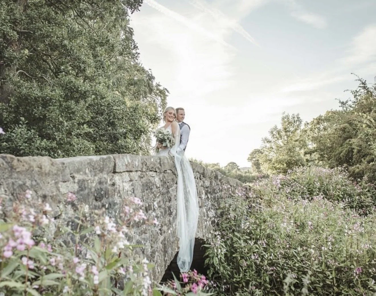 A wedding couple on a stone wall surrounded by greenery and pink flowers, with a bright sky backdrop.